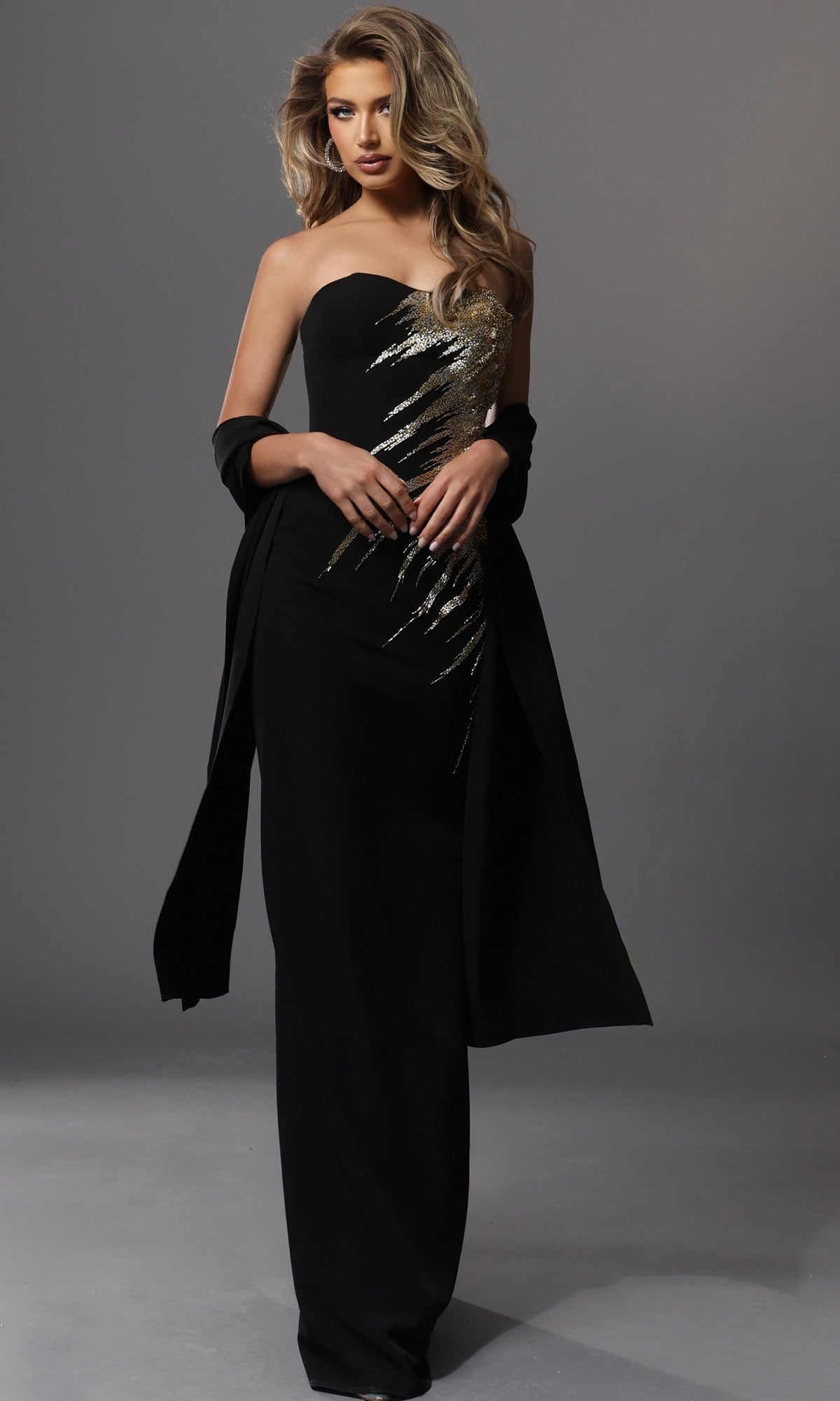 A woman stands against a plain gray background, wearing the Jovani 51900 long formal dress—a fitted black sheath silhouette with a strapless neckline, embellished with gold sequin embroidery—and a matching black shawl.